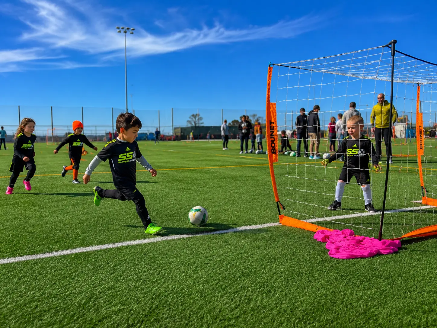 Young Super Soccer Stars player shooting into a small goal while a teammate keeps net on a sunny turf field